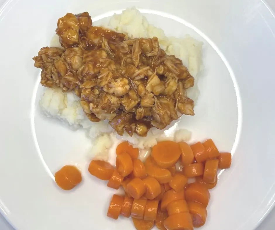 white plate with buttered carrots and bbq chicken on top of mashed potatoes