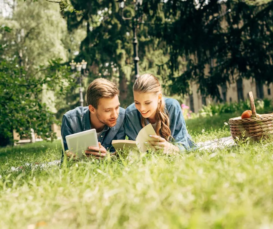 couple smiling, each reading something at a picnic and laying on the blanket