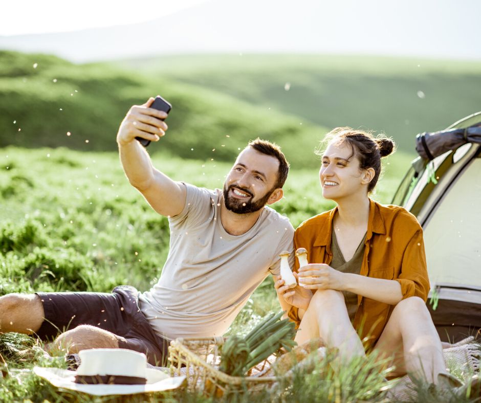 couple picnicking in meadow taking selfie, in front of a tent