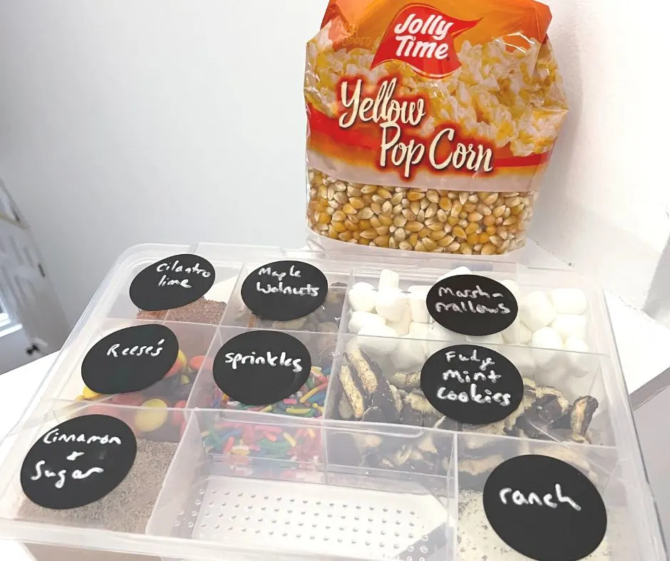 bag of popcorn behind a tacklebox filled with different popcorn toppings and black labels with chalk writing
