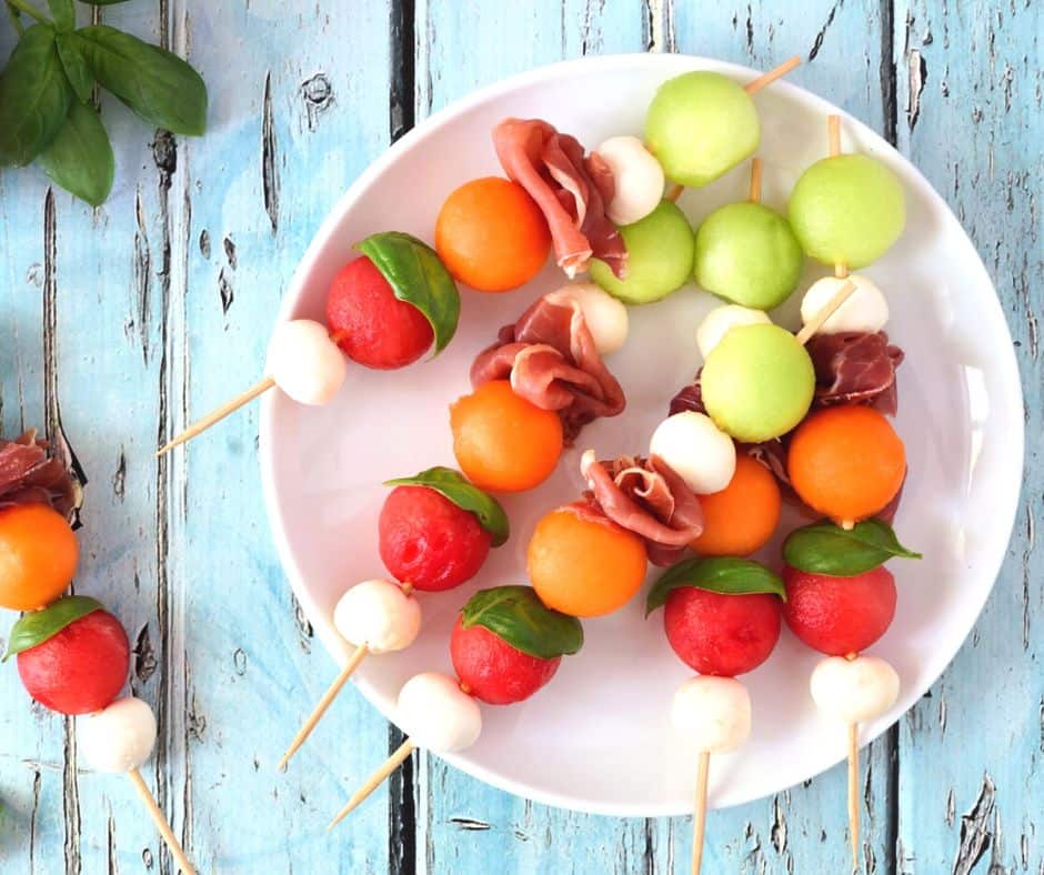 white plate of melon, prosciutto skewers on light blue wooden table