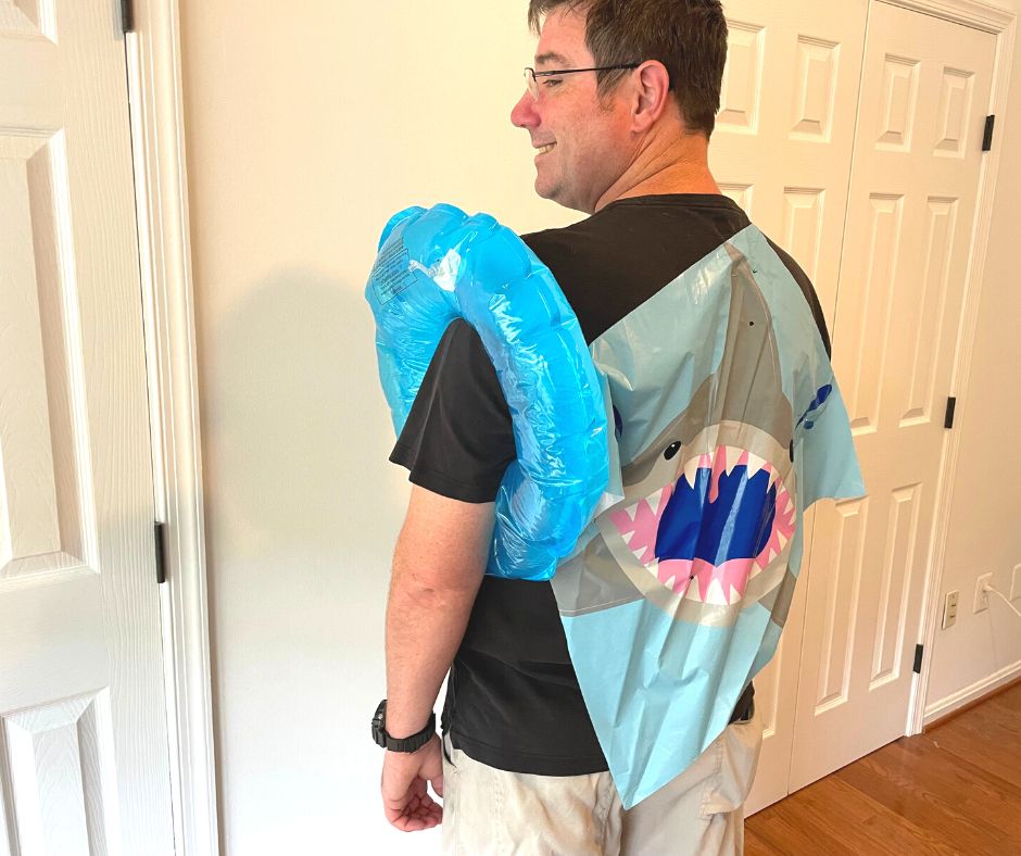 man in black shirt with small, blown-up inflatable blue swimming device on arm and big shark kite on back