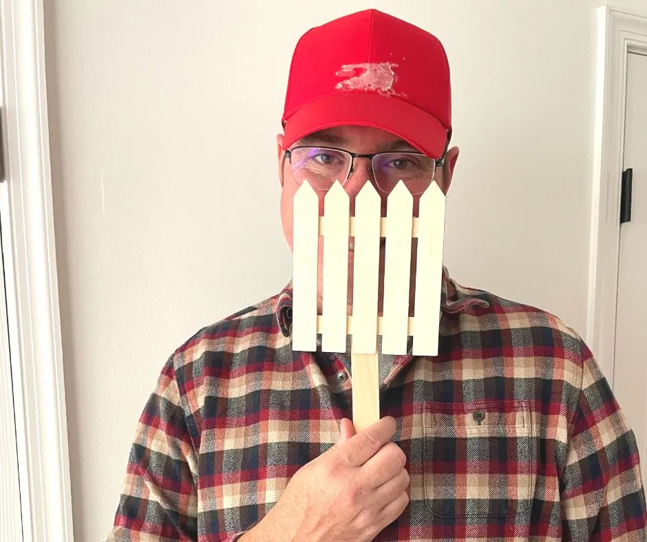 man with glasses in plaid shirt and red hat, holding up small wooden fence