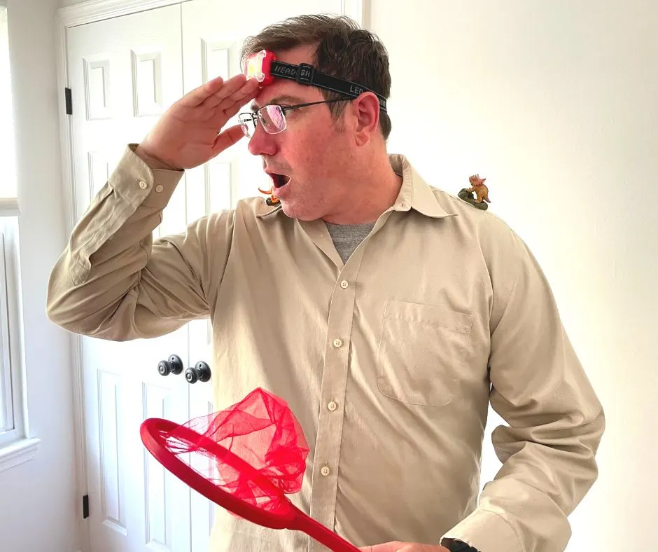 man looking surprised with red net, tan shirt, headlamp, and two small dinosaur toys on shoulders