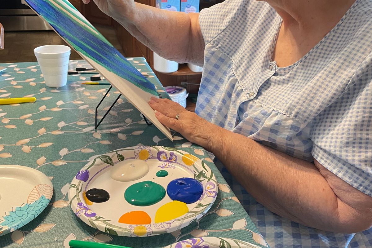 woman painting using all Dollar Tree supplies with plate of paint colors, brushes, easel, canvas, etc. 