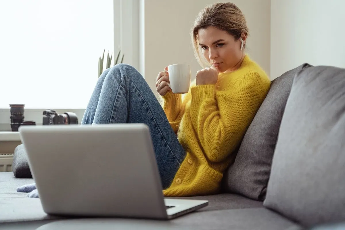 woman in yellow sweater looking pensively at laptop with earbuds listening to crime podcast on couch