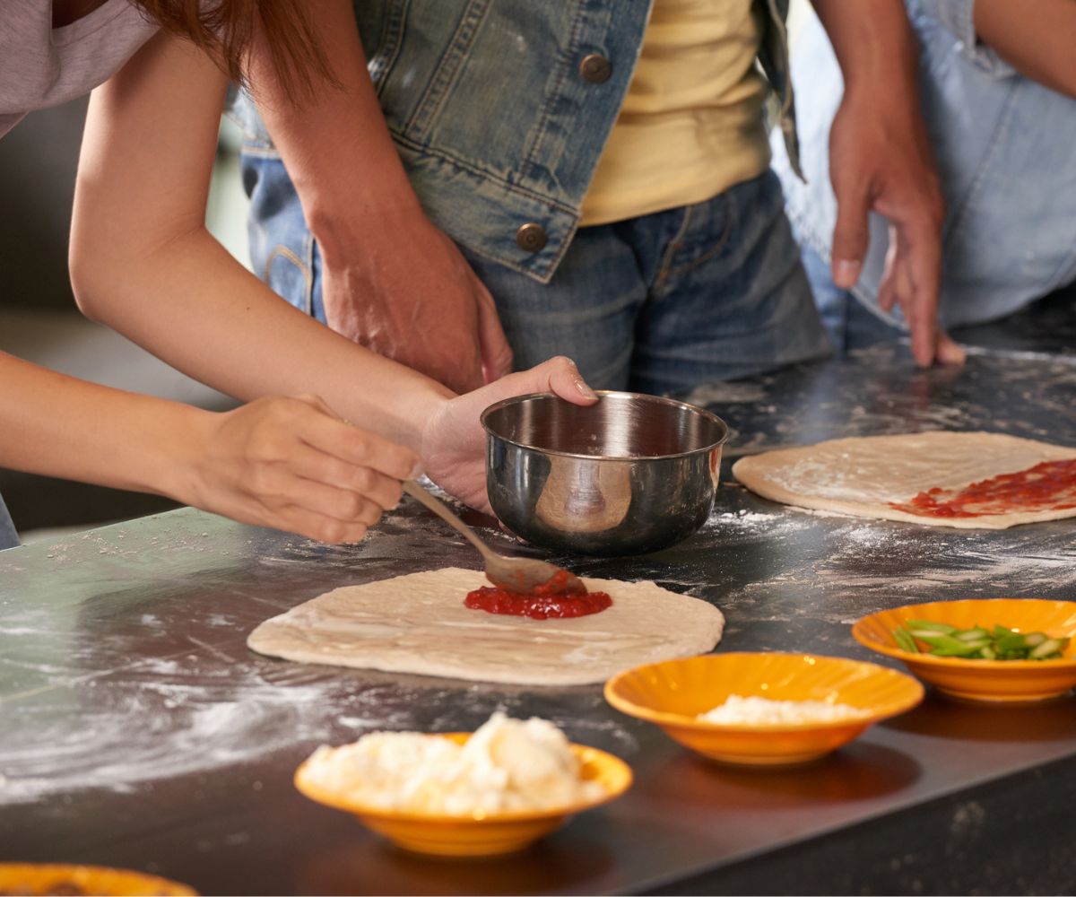 group of people at kitchen bar spreading pizza sauce and putting toppings on homemade pizza dough