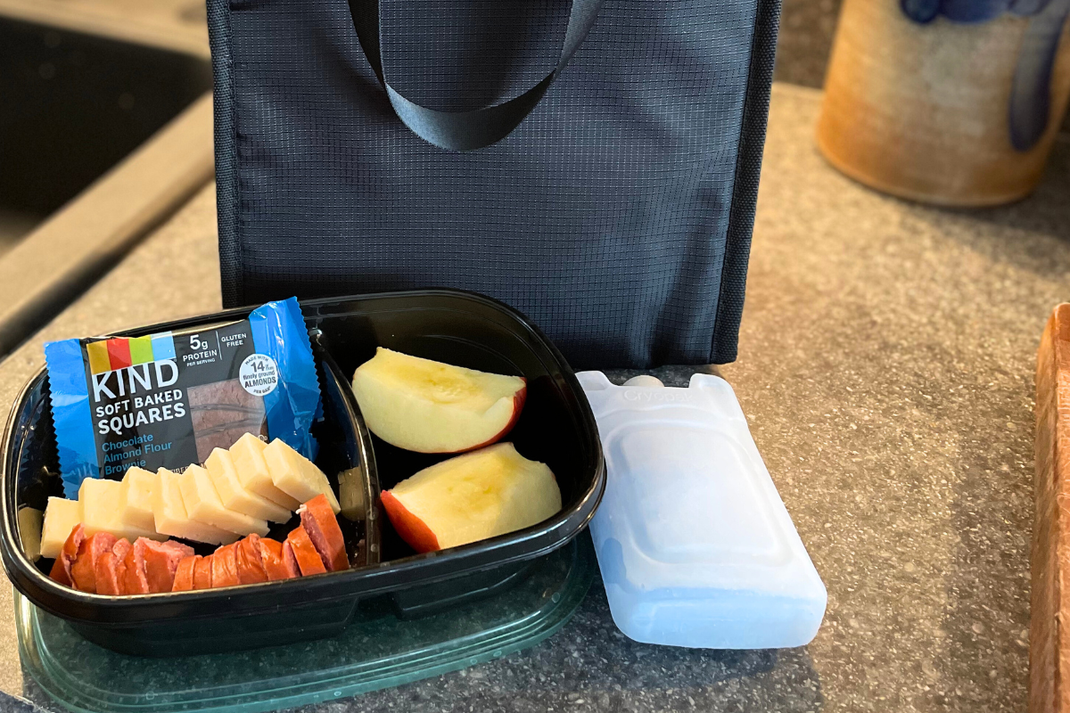 black lunch bag with black lunch container of sausage, cheese, apples, etc. on countertop