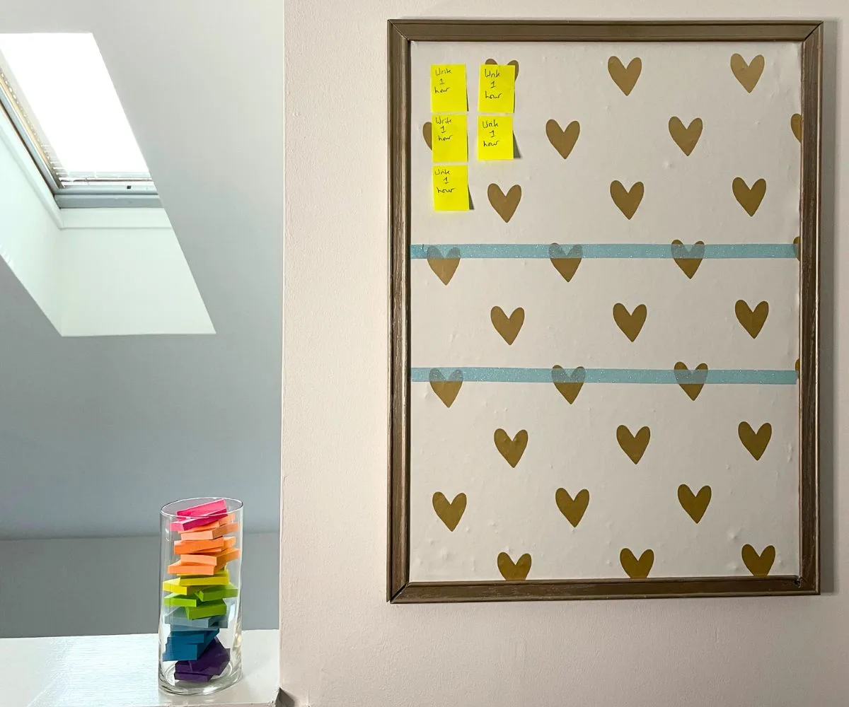 vertical bulletin board with gold hearts wallpaper on it, and small post it notes saying "Write 1 Hour" on each, next to vase filled with small post-it notes