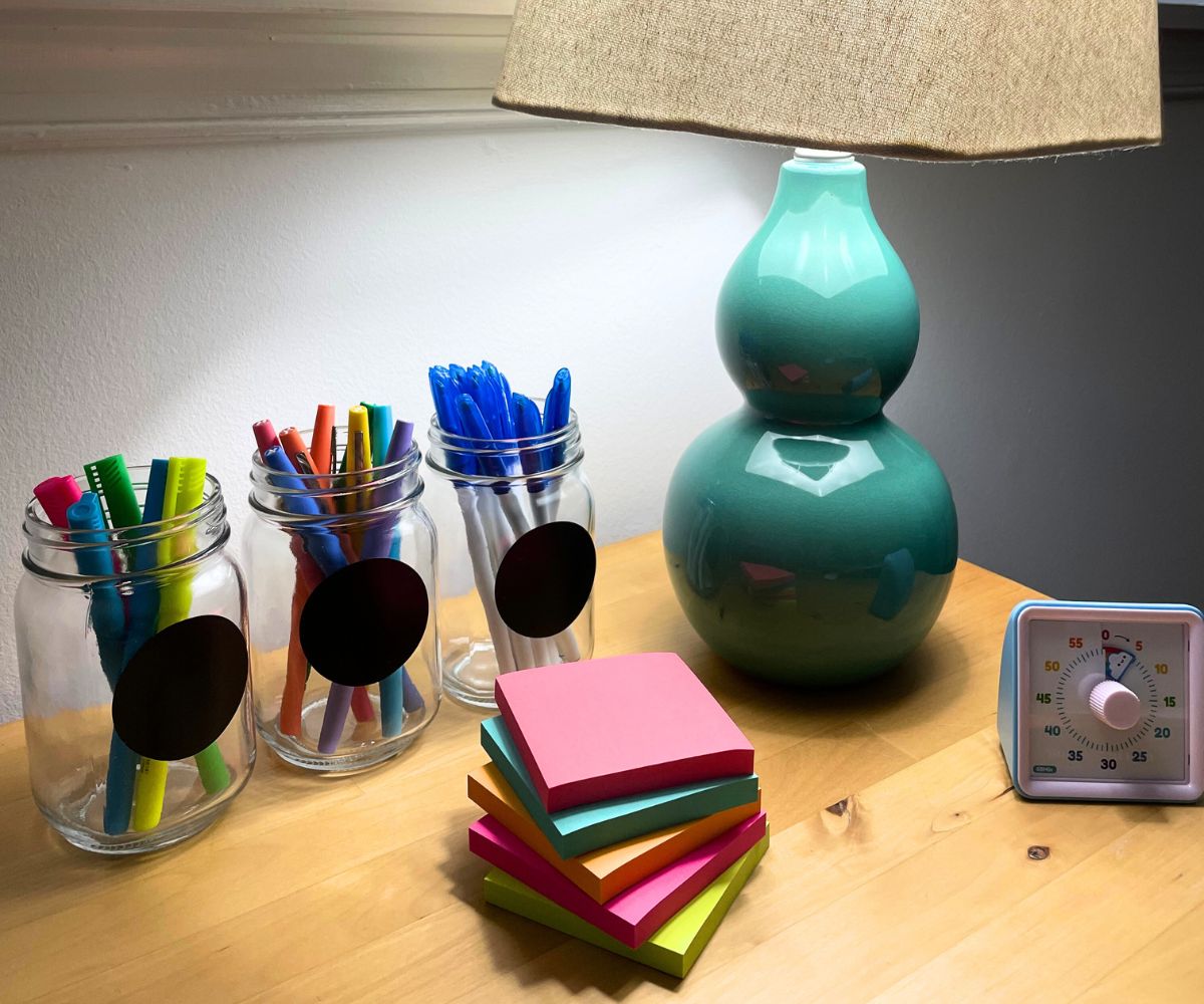 three mason jars with circle chalkboard sticker, with pens, highlighters, and markers in front of turquoise lamp and post-it notes stack