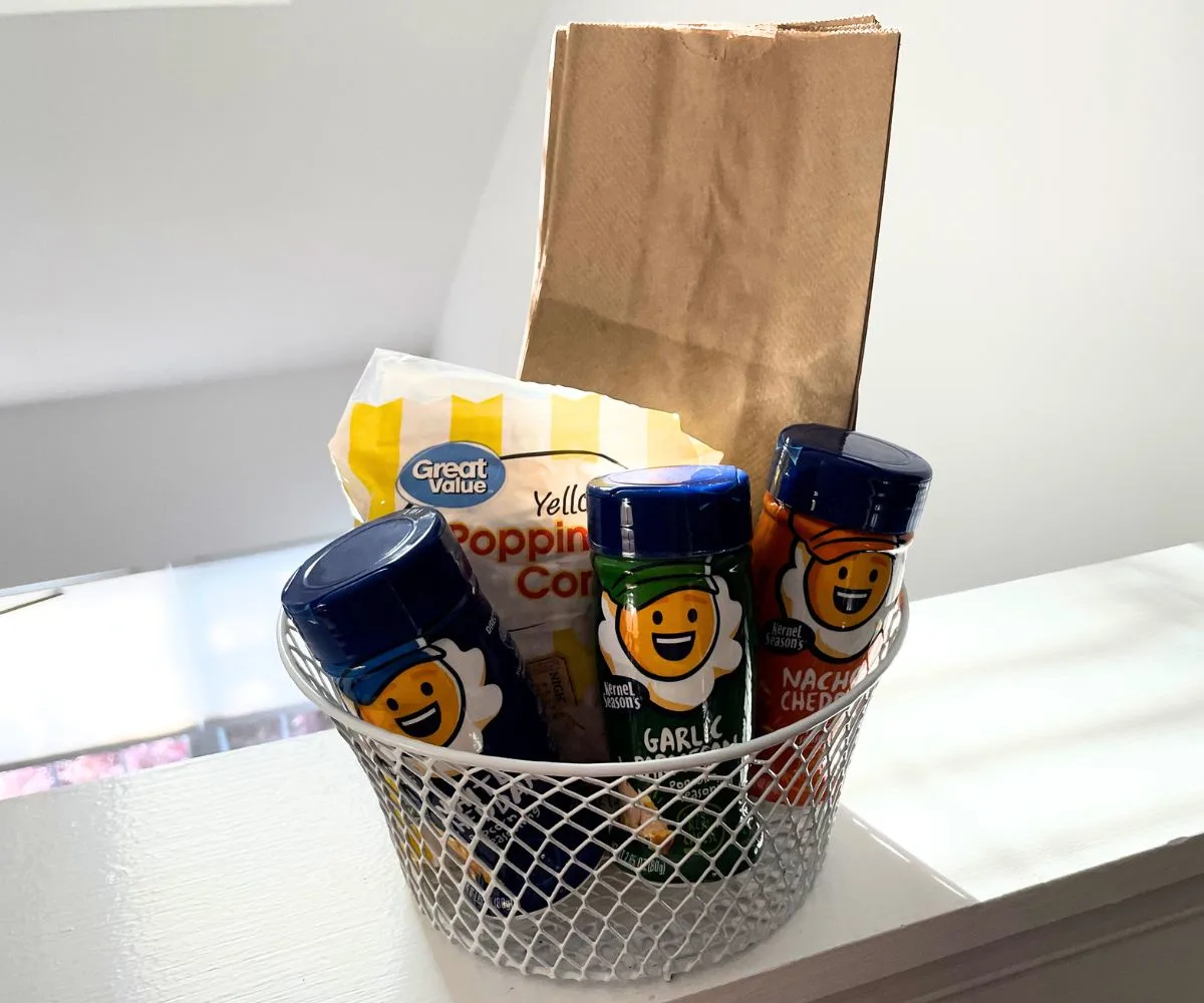 white wire basket filled with popcorn kernel bag, lunch bags, and three popcorn seasoning flavors
