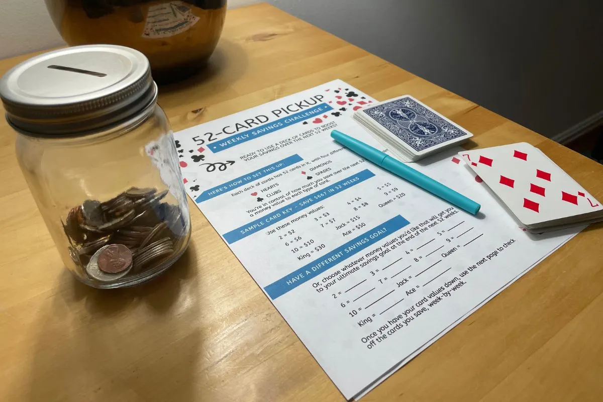 mason jar 1/3 filled with coins with coin-slotted lid, 52-week printable savings challenge, and deck of cards on wooden table