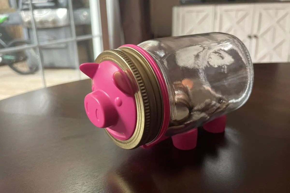 mason jar on its side as body of pink pig, pink pig head with ears and stout feet on bottom, on dark table