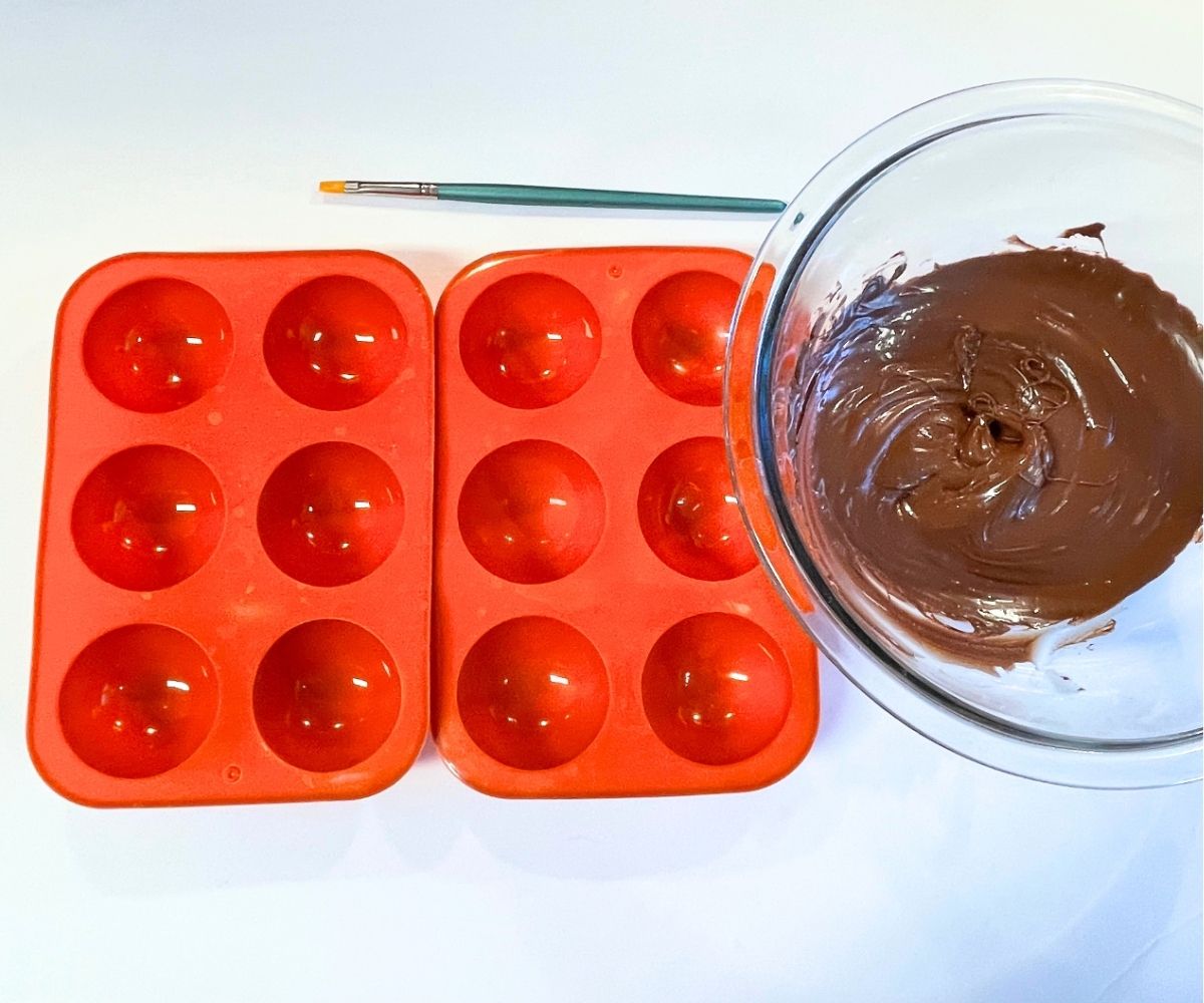 melted chocolate in bowl next to two silicon hot cocoa bombshell molds and a small paintbrush