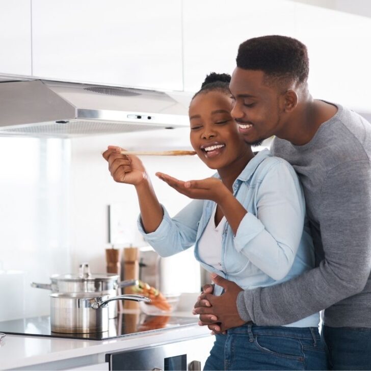 couple cooking in kitchen together, husband tasting from pot