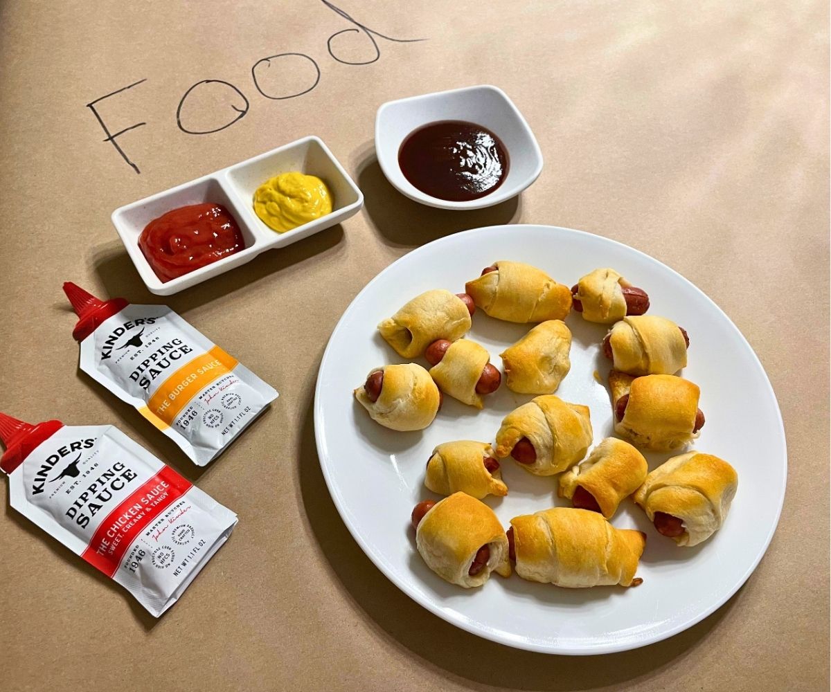 plate full of pigs in a blanket, with lots of sauces under "Food" written in marker on butcher paper