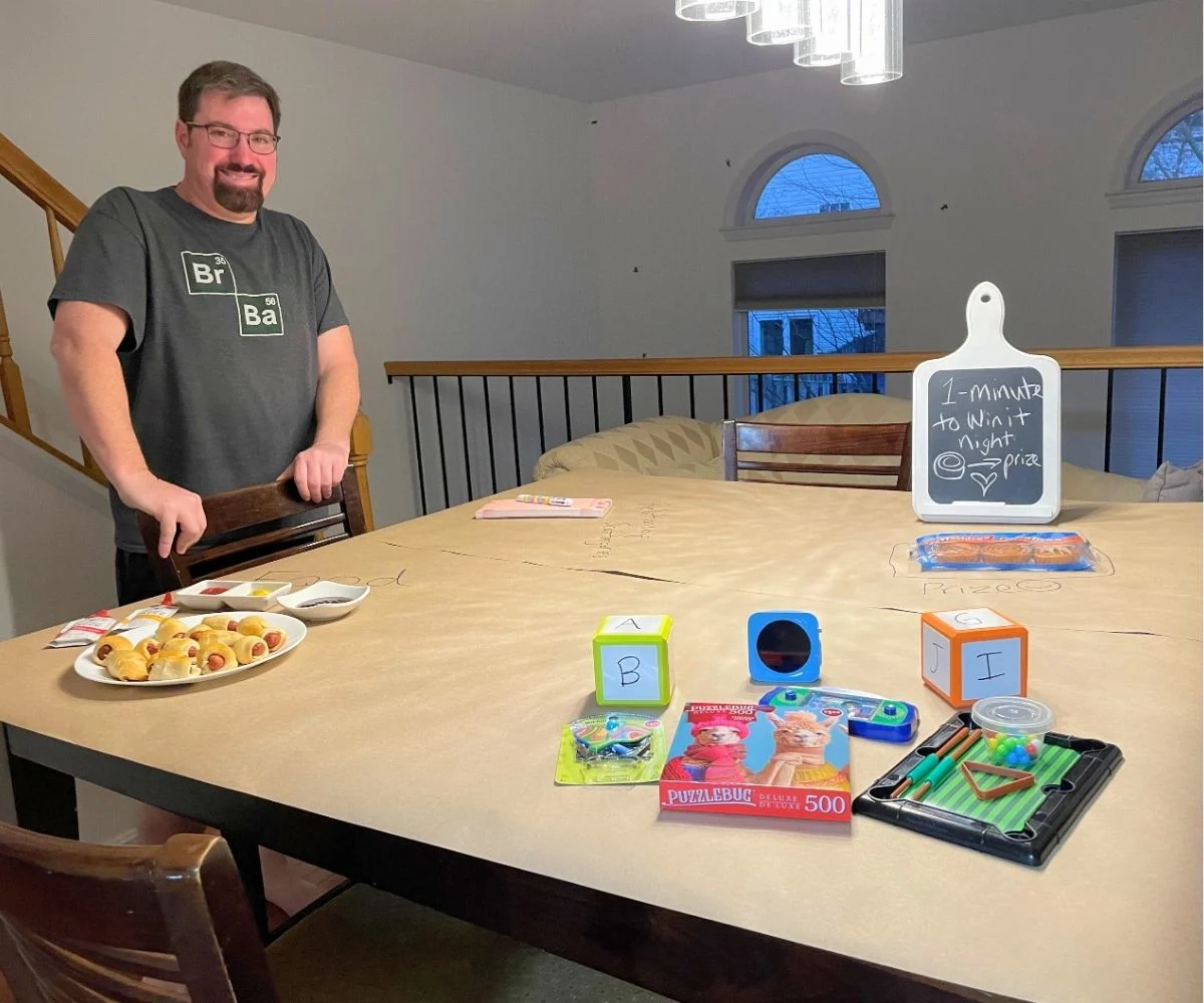 husband smiling, surprised, looking over whole dining room table covered in butcher paper with games and such