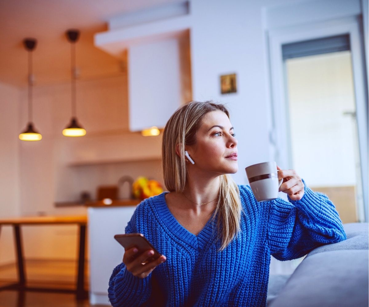 woman in blue sweater looking pensively out of window and listening to crime podcast