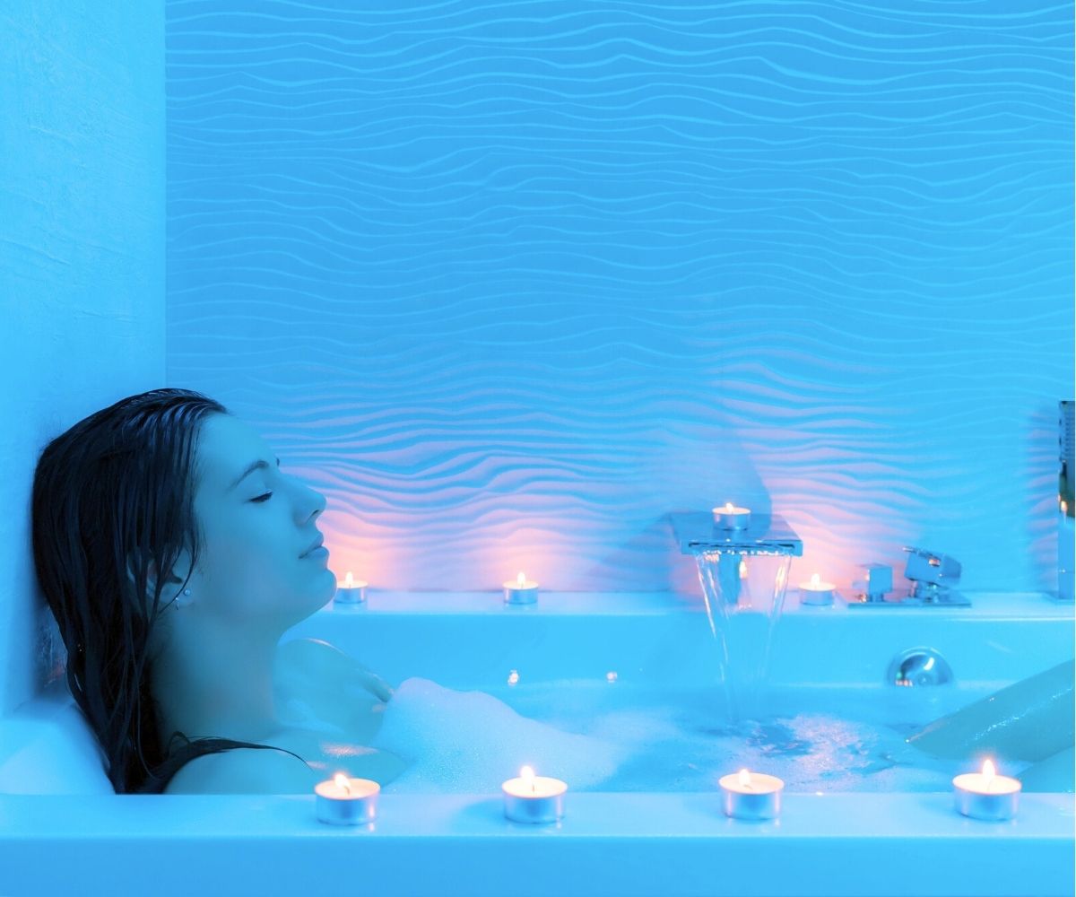 woman with eyes closed and candles all around in blue-lit bathtub area