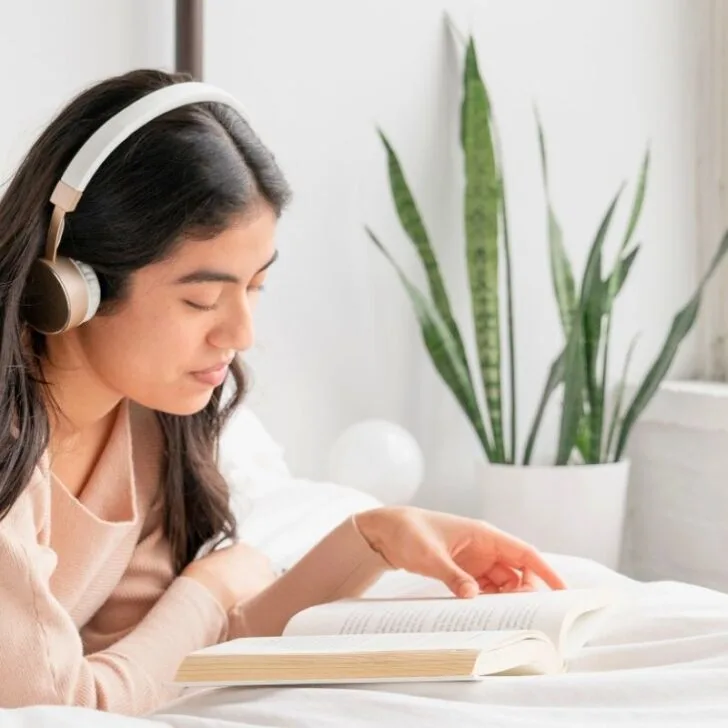 woman with headphones reading book in a white themed room with plant