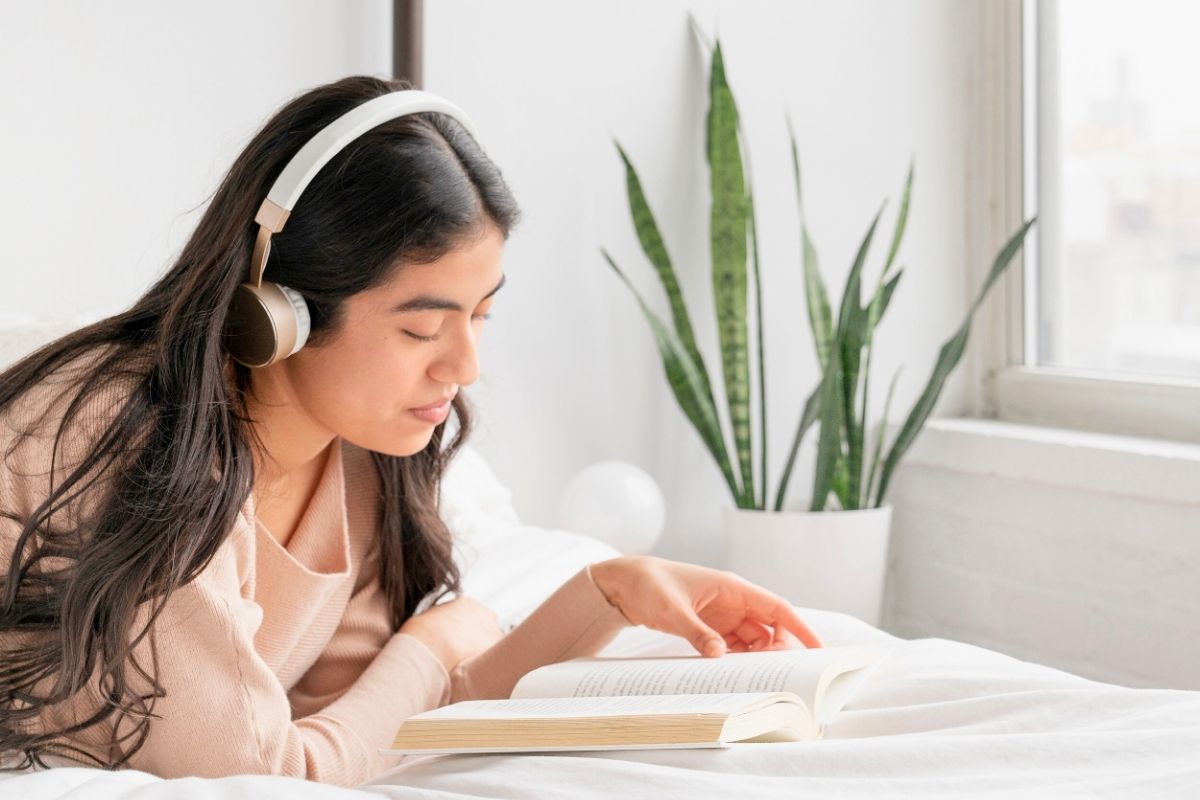 woman with headphones reading book in a white themed room with plant