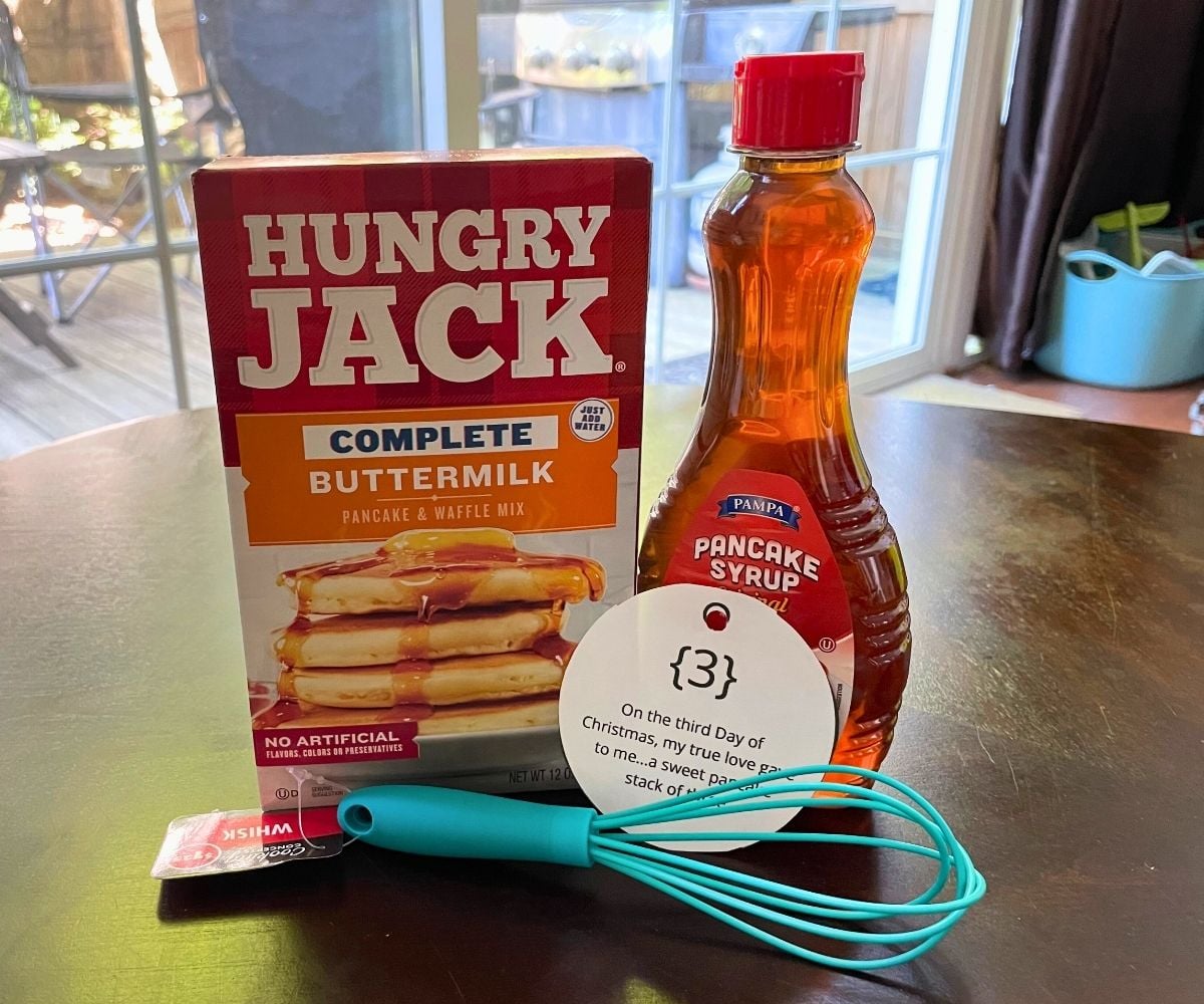 box of buttermilk pancakes, small teal whisk, and bottle of pancake syrup on dark table