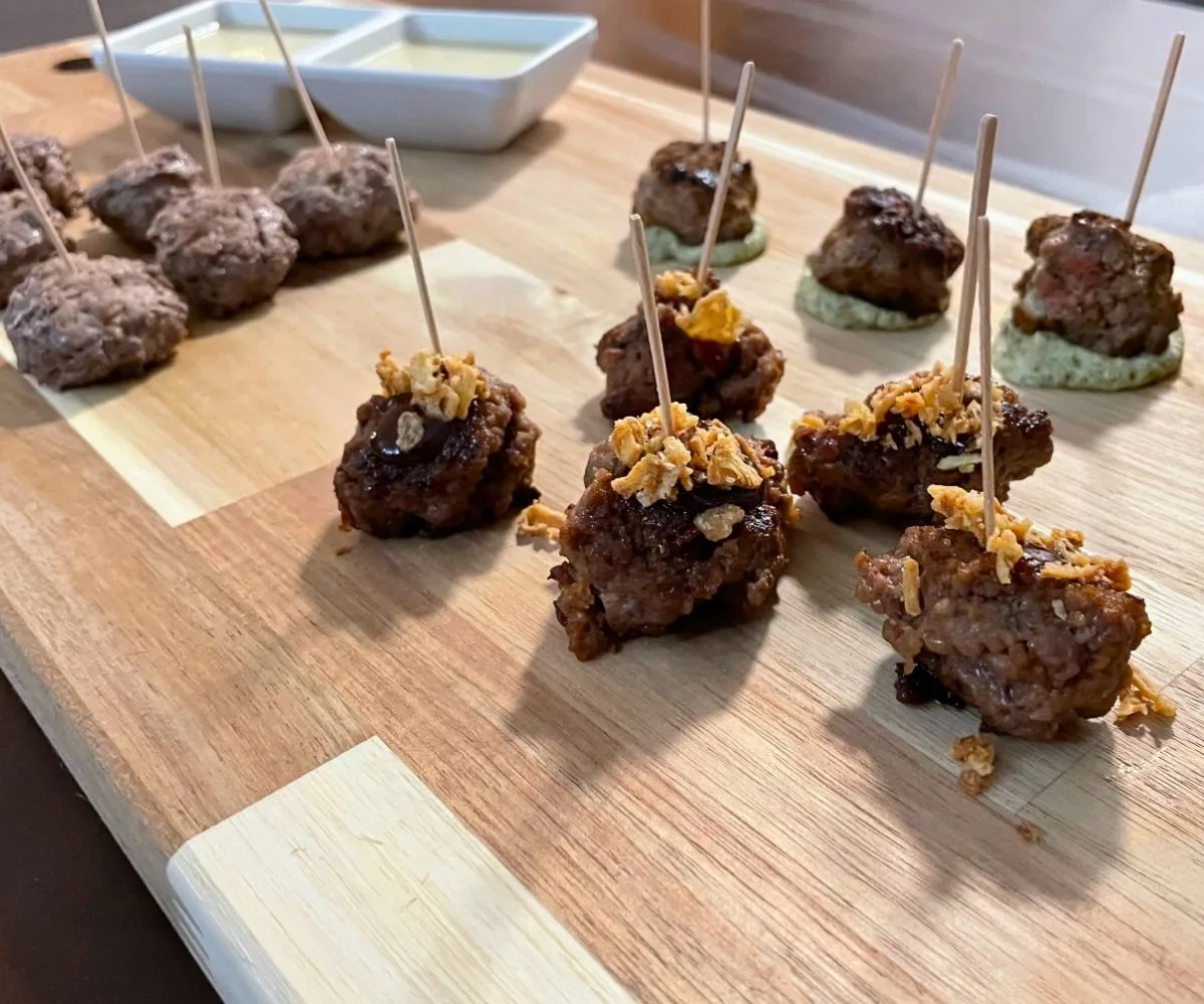 light wooden board filled with three different kinds of meatballs, with toothpicks out of each one, and two sauce dishes