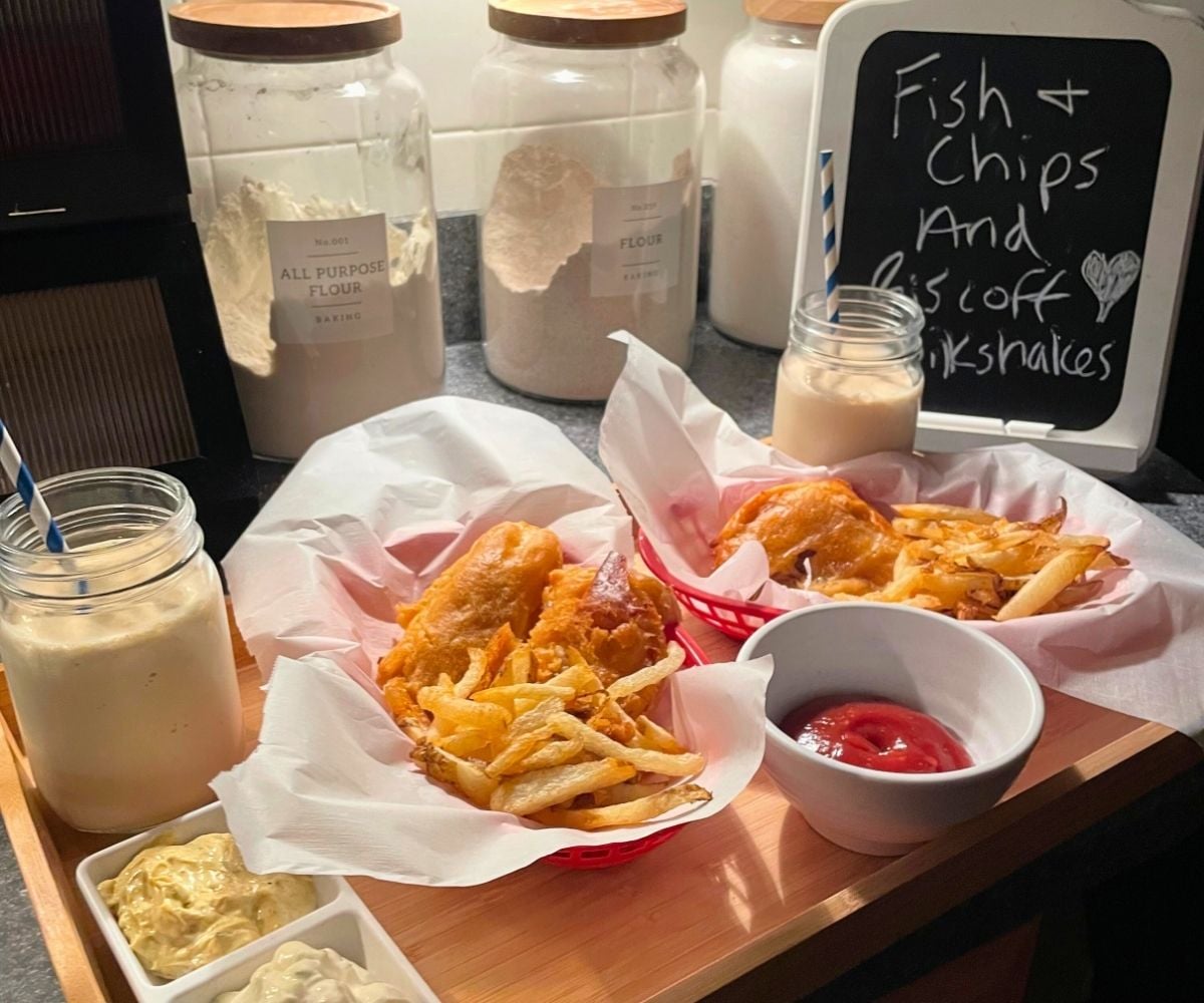 light wooden tray filled with red baskets of fried fish and chips, two biscoff milkshakes with blue and white striped straws, sauces, and a chalkboard detailing the meal