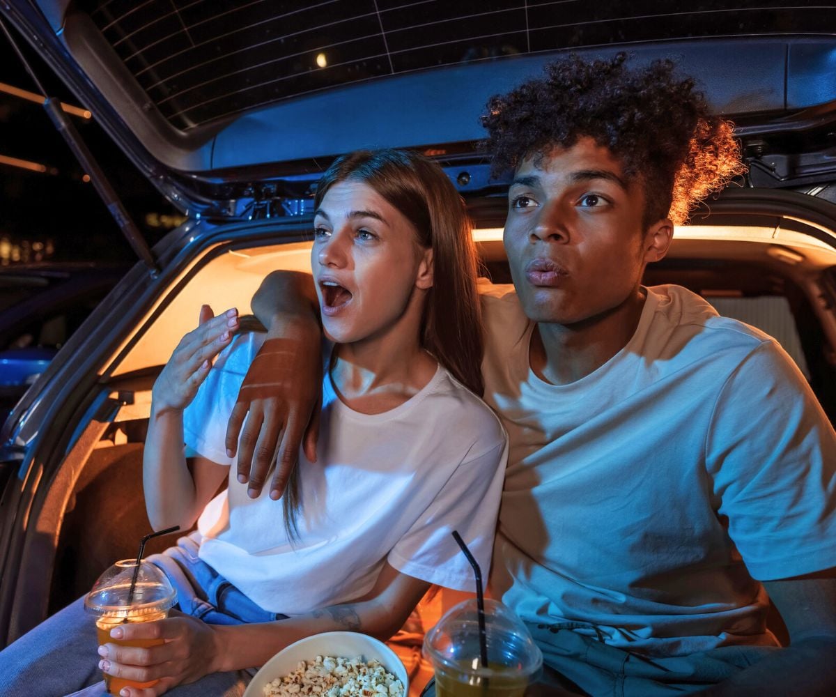 young night in back of car with popcorn, looking shocked at drive in movie screen