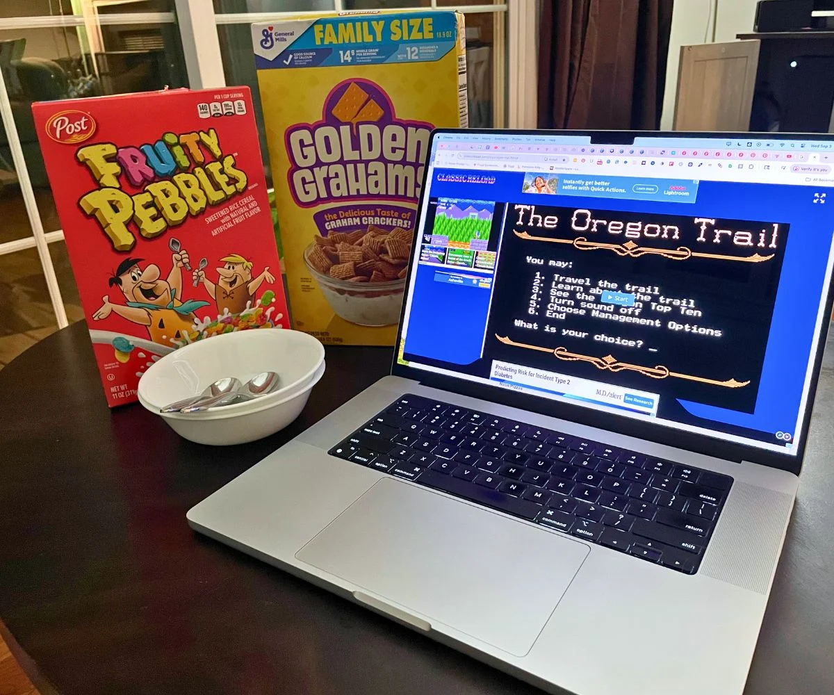 laptop screen showing Oregon Trail original game, next to Fruity Pebbles, Golden Grahams, and two bowls with spoons