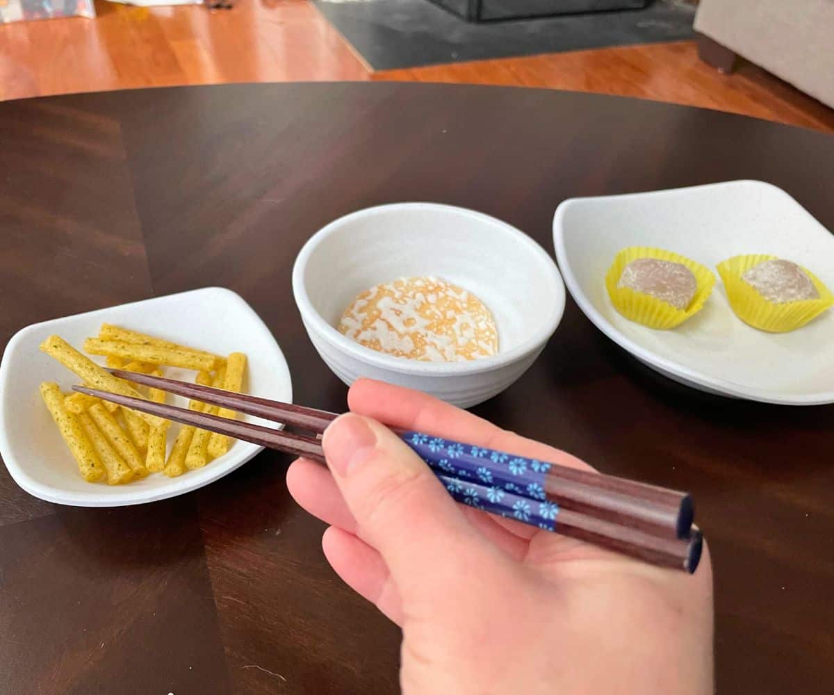 Woman using chopsticks to eat three different kinds of international snacks on dark table
