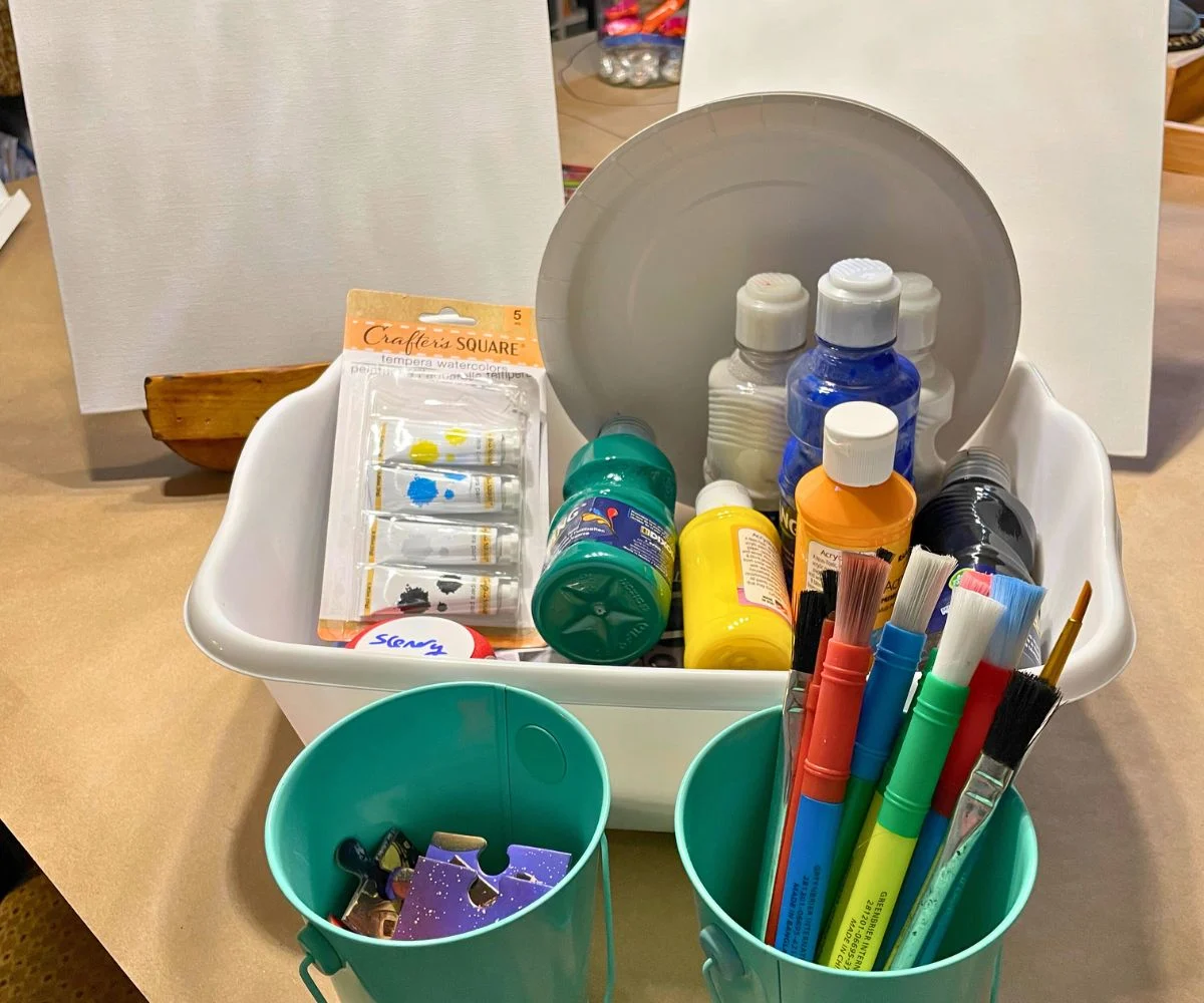 bucket of puzzle pieces, bucket of paintbrushes, and bucket of paints with canvas behind them