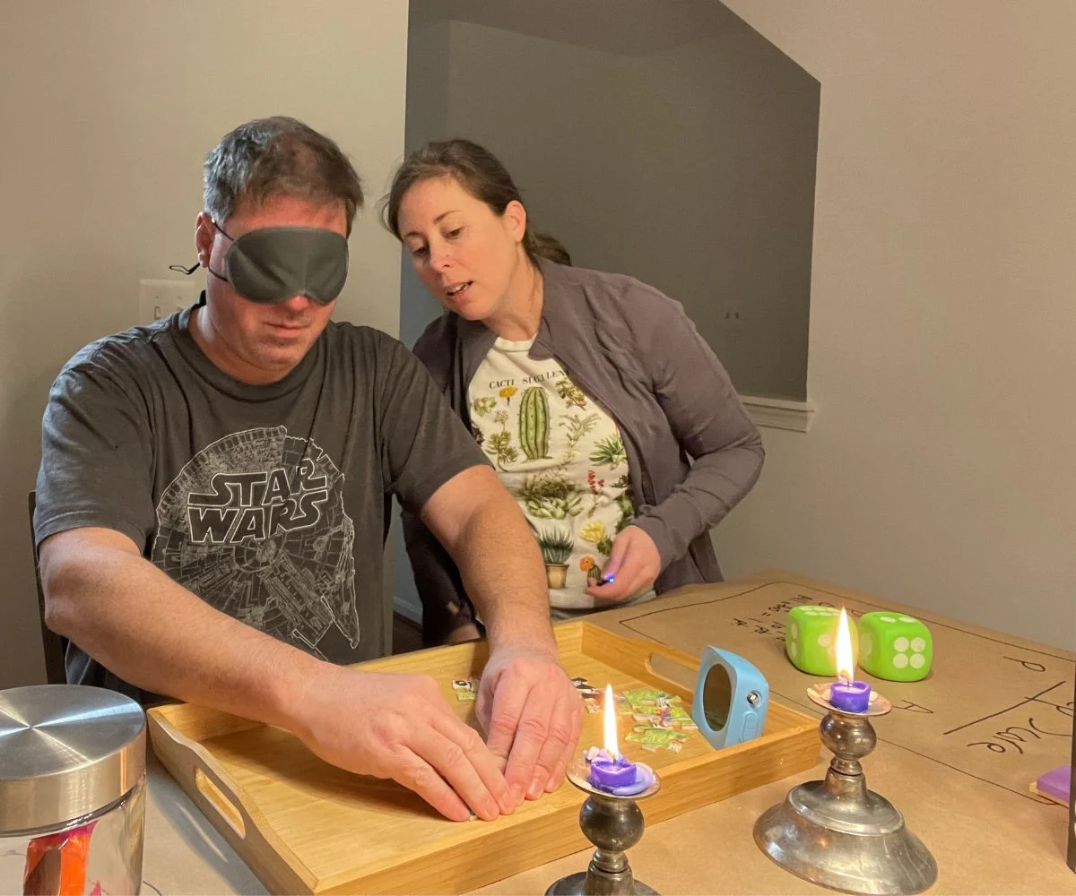 husband blindfolded with hands on puzzle pieces in tray, wife giving him directions