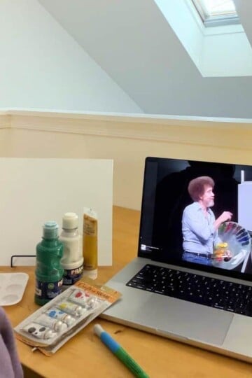 author looking at Bob Ross on computer screen, with painting supplies in front of her