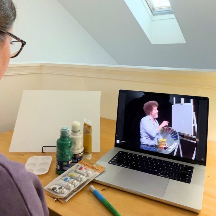 author looking at Bob Ross on computer screen, with painting supplies in front of her