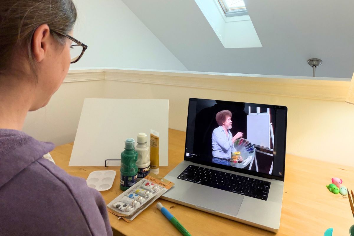 author looking at Bob Ross on computer screen, with painting supplies in front of her