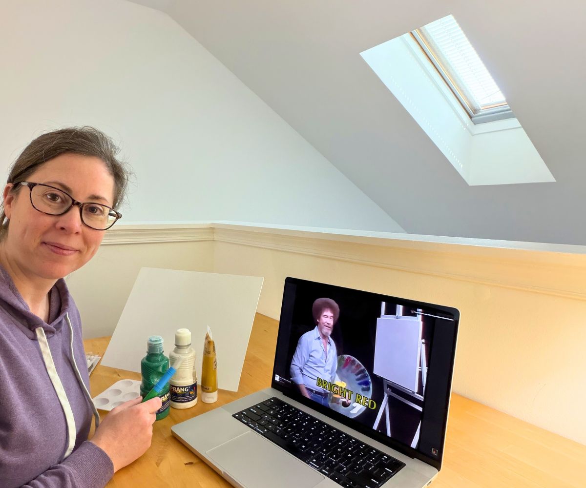author smiling at camera with Bob Ross on computer screen and painting supplies in front of her