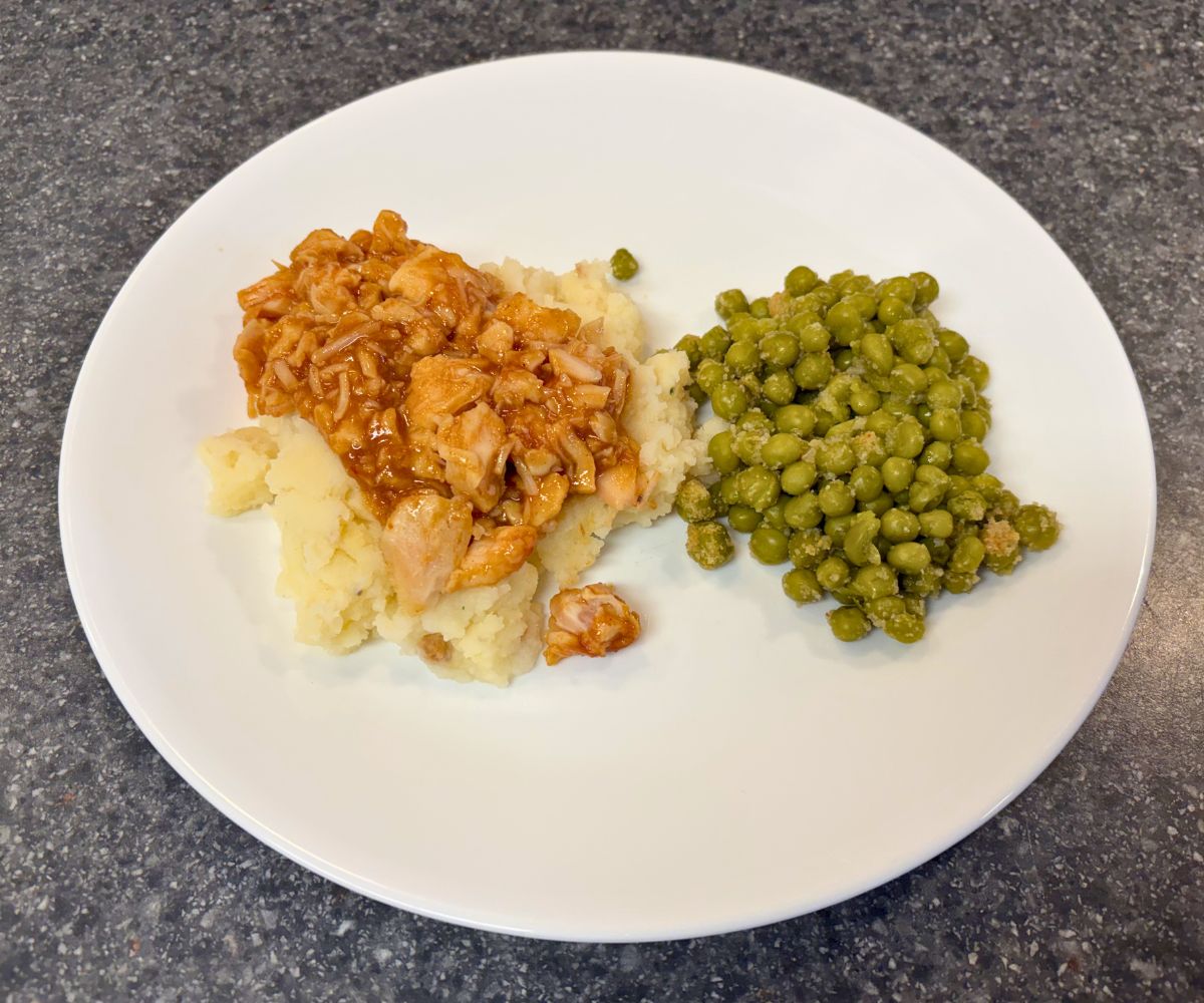 pile of BBQ chicken on pillow of mashed potatoes, next to a cup of sweet peas, on a white plate